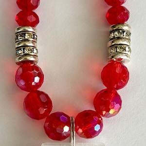 May include: A red beaded necklace with a pendant. The necklace features faceted red beads, silver-colored spacers, and a rectangular pendant with the word "JOKER" and a green design.