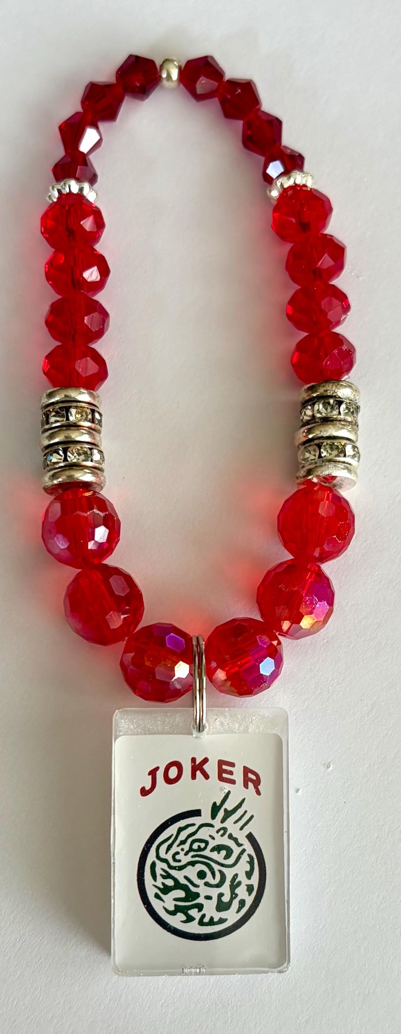 May include: A red beaded necklace with a pendant. The necklace features faceted red beads, silver-colored spacers, and a rectangular pendant with the word "JOKER" and a green design.