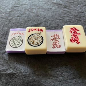 May include: Four ivory-colored Mahjong tiles with purple edges. Two tiles read "JOKER" with dragon graphics, one with a red dragon, and one with a dragon in a circle. The tiles are arranged on a gray surface.