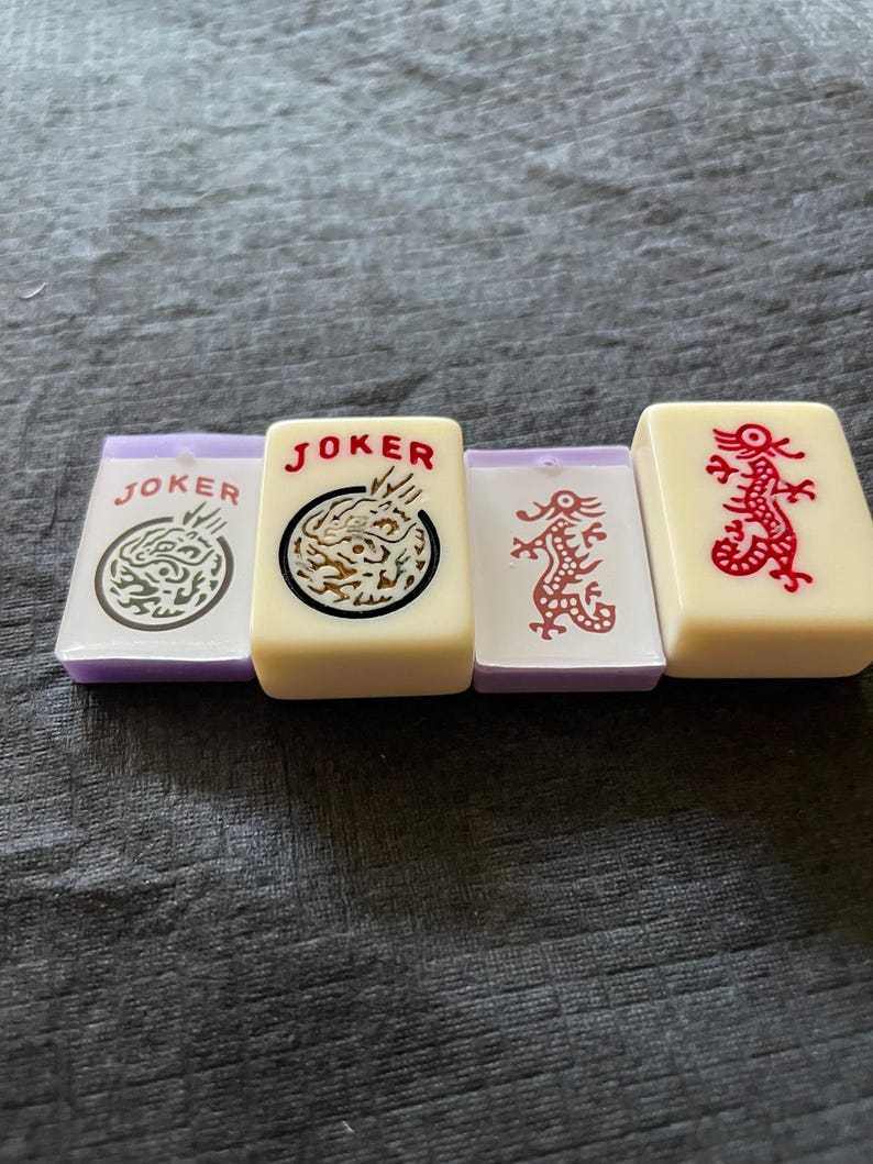 May include: Four ivory-colored Mahjong tiles with purple edges. Two tiles read "JOKER" with dragon graphics, one with a red dragon, and one with a dragon in a circle. The tiles are arranged on a gray surface.