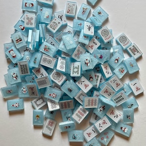 May include: A collection of light blue Mahjong tiles with various symbols, numbers, and festive holiday designs. The tiles feature red, white, and black details, including snowmen and snowflakes. A fun game for the holidays.