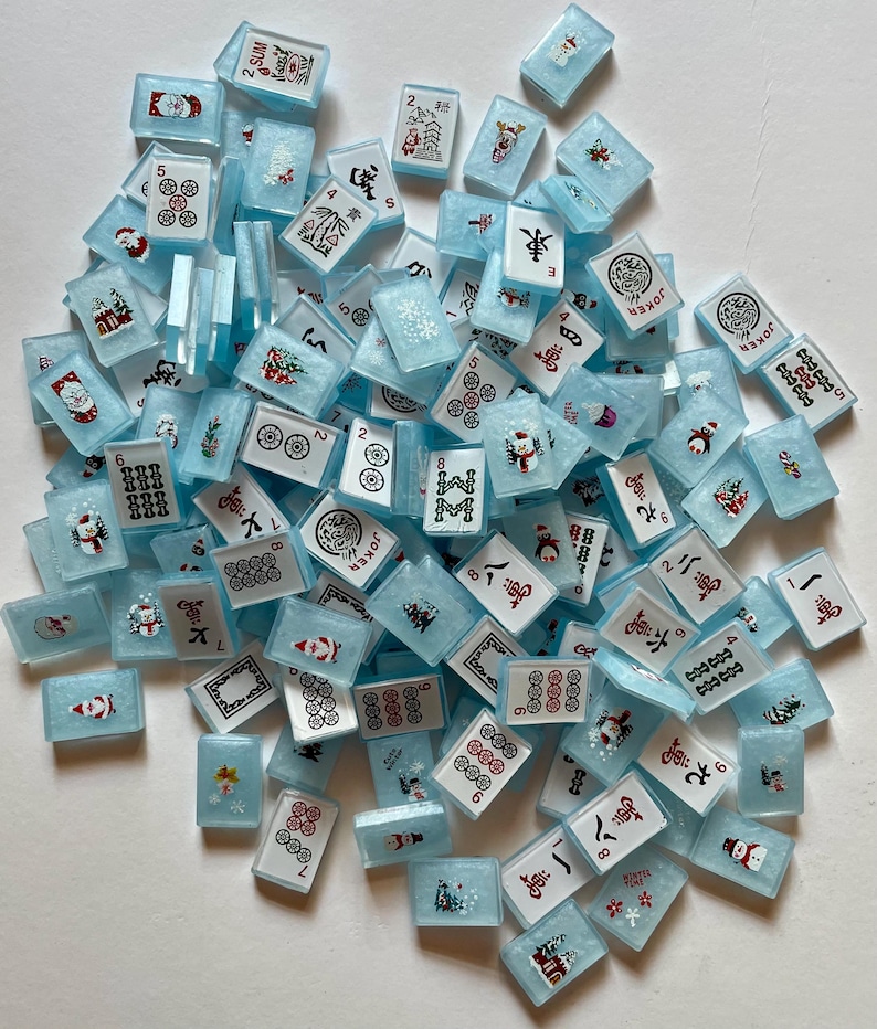 May include: A collection of light blue Mahjong tiles with various symbols, numbers, and festive holiday designs. The tiles feature red, white, and black details, including snowmen and snowflakes. A fun game for the holidays.