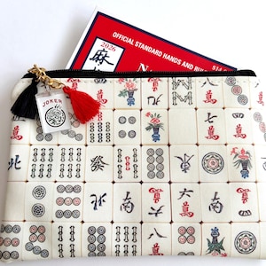 May include: A rectangular cream-colored Mahjong tile print zippered pouch with a black zipper and gold-colored chain. A red tassel and a black tassel hang from the zipper. A small clear charm with the word "JOKER" is attached. A red and white book is inside the pouch.