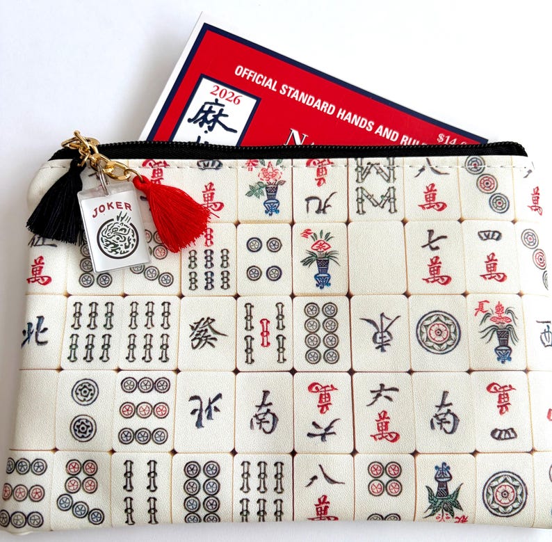 May include: A rectangular cream-colored Mahjong tile print zippered pouch with a black zipper and gold-colored chain. A red tassel and a black tassel hang from the zipper. A small clear charm with the word "JOKER" is attached. A red and white book is inside the pouch.