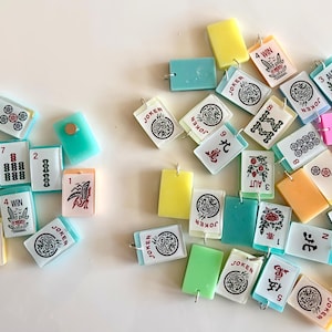 May include: A collection of colorful Mahjong tiles scattered on a white surface. The tiles feature various symbols, numbers, and Chinese characters, including "Joker" and "Win". The tiles are in shades of green, yellow, pink, and white.