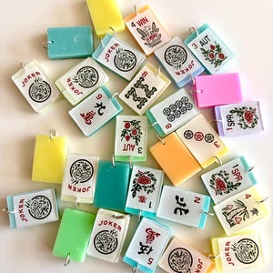 May include: A collection of colorful, rectangular Mahjong tile charms with silver-tone loops. The tiles feature various symbols, numbers, and the word "JOKER" in black and red. Some tiles are solid colors like yellow, green, and pink.