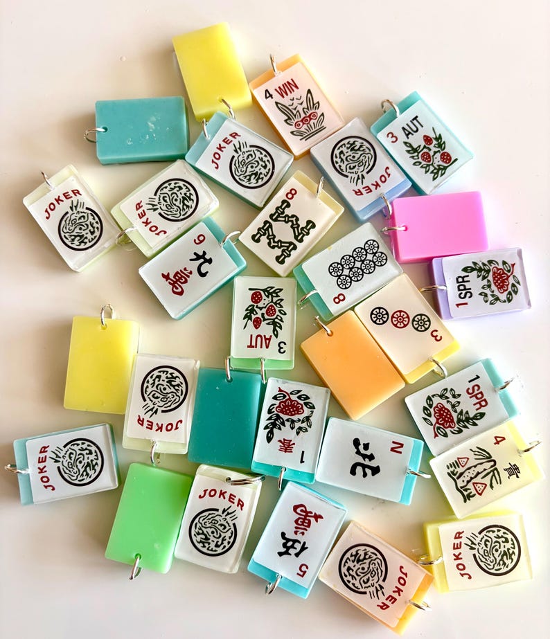 May include: A collection of colorful, rectangular Mahjong tile charms with silver-tone loops. The tiles feature various symbols, numbers, and the word "JOKER" in black and red. Some tiles are solid colors like yellow, green, and pink.