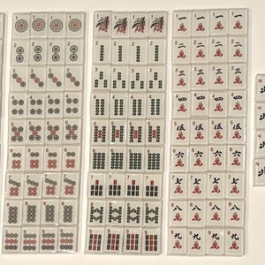 May include: A collection of white Mahjong tiles with red and black symbols and characters. The tiles are arranged in several stacks, displaying various suits, numbers, and directional markers. The set includes Jokers and flower tiles.