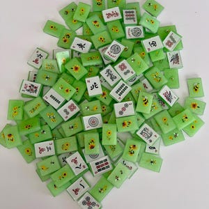 May include: A collection of green Mahjong tiles with various designs, including sunflowers, bees, and Chinese characters. The tiles are rectangular and have white faces with black, red, and yellow details. Some tiles display numbers and symbols.