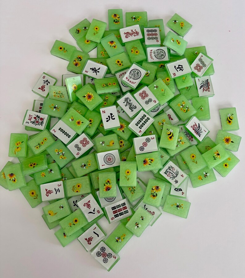May include: A collection of green Mahjong tiles with various designs, including sunflowers, bees, and Chinese characters. The tiles are rectangular and have white faces with black, red, and yellow details. Some tiles display numbers and symbols.