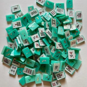 May include: A collection of turquoise Mahjong tiles scattered on a white surface. The tiles feature various symbols, numbers, and Chinese characters in black and red. Some tiles have decorative fish and floral designs.