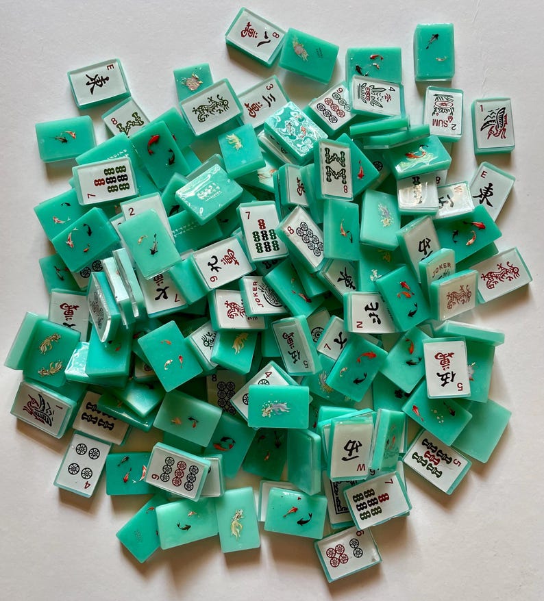May include: A collection of turquoise Mahjong tiles scattered on a white surface. The tiles feature various symbols, numbers, and Chinese characters in black and red. Some tiles have decorative fish and floral designs.
