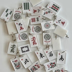 May include: A collection of small, rectangular Mahjong tile keychains. The keychains are white with various symbols, numbers, and Chinese characters in red, green, and black. Some keychains have the word "JOKER" on them.