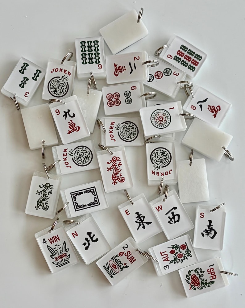 May include: A collection of small, rectangular Mahjong tile keychains. The keychains are white with various symbols, numbers, and Chinese characters in red, green, and black. Some keychains have the word "JOKER" on them.