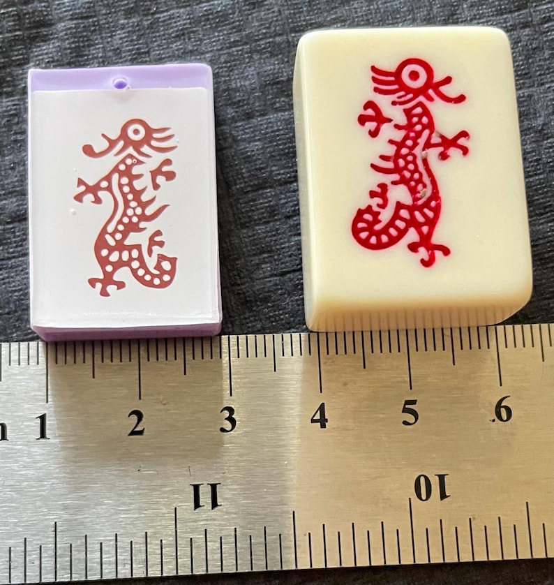 May include: Two Mahjong tiles. One is a rectangular, off-white tile with a red dragon design. The other is a smaller, rectangular tile with a purple frame and a white face with a red dragon design. A ruler is in the background.