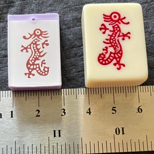 May include: Two Mahjong tiles. One is a light purple and white tile with a red dragon design. The other is an off-white tile with a red dragon design. A ruler is in the background, showing measurements in inches.
