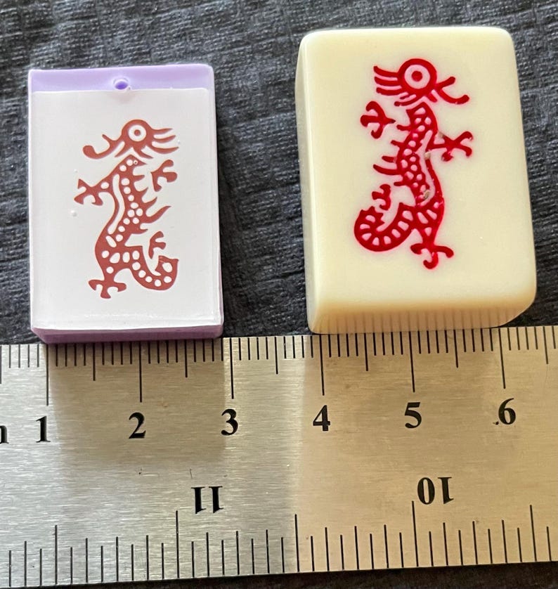 May include: Two Mahjong tiles. One is a light purple and white tile with a red dragon design. The other is an off-white tile with a red dragon design. A ruler is in the background, showing measurements in inches.