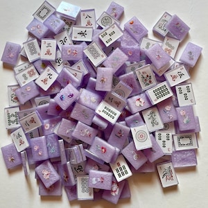 May include: A collection of lavender-colored Mahjong tiles with various symbols and numbers in black and red. Some tiles feature floral designs. The tiles are scattered, showing a variety of suits and characters.