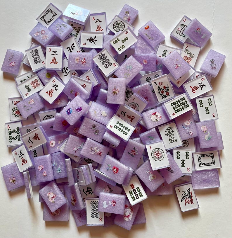 May include: A collection of lavender-colored Mahjong tiles with various symbols and numbers in black and red. Some tiles feature floral designs. The tiles are scattered, showing a variety of suits and characters.