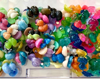 May include: A collection of small, colorful duck-shaped resin charms in a clear plastic container. The charms feature various translucent, opaque, and glittery finishes in shades of blue, green, pink, orange, yellow, purple, black, and white.