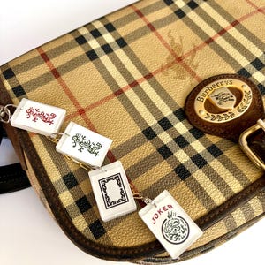 May include: Tan and black plaid Burberry shoulder bag with a brown leather strap and gold buckle. The bag features a gold emblem with the Burberry logo. Attached are four white rectangular charms with various designs and the word "JOKER".