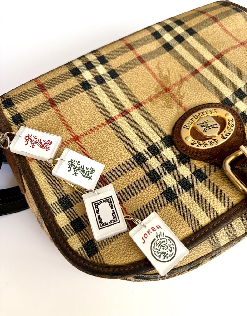 May include: Tan and black plaid Burberry shoulder bag with a brown leather strap and gold buckle. The bag features a gold emblem with the Burberry logo. Attached are four white rectangular charms with various designs and the word "JOKER".