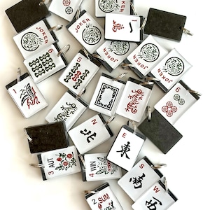 May include: A collection of white and black Mahjong tile keychains with various symbols, numbers, and Chinese characters. Some tiles feature the word "JOKER" and others have red and green designs. Each keychain has a silver ring.