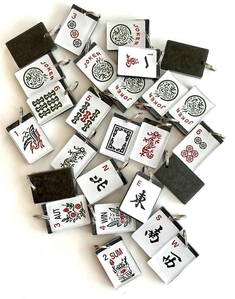 May include: A collection of white and black Mahjong tile keychains with various symbols, numbers, and Chinese characters. Some tiles feature the word "JOKER" and others have red and green designs. Each keychain has a silver ring.
