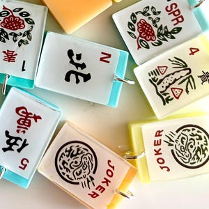 May include: A collection of colorful, rectangular Mahjong tile charms. Each charm features unique symbols, numbers, and Chinese characters in red and black, with a silver loop for attachment. The tiles have a white background with light blue or yellow edges.
