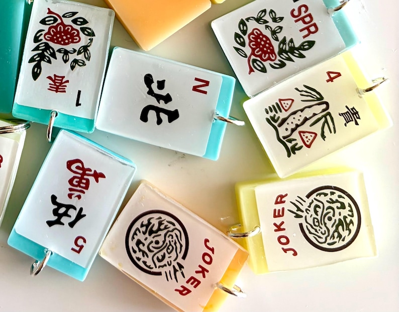 May include: A collection of colorful, rectangular Mahjong tile charms. Each charm features unique symbols, numbers, and Chinese characters in red and black, with a silver loop for attachment. The tiles have a white background with light blue or yellow edges.