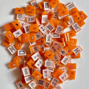 May include: A collection of orange and white Mahjong tiles scattered across a white surface. The tiles feature various symbols, numbers, and characters in black and red. Some tiles have illustrations of flowers, cats, and other designs. The game pieces are rectangular.
