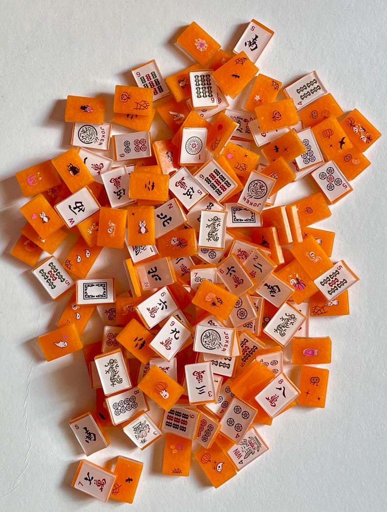 May include: A collection of orange and white Mahjong tiles scattered across a white surface. The tiles feature various symbols, numbers, and characters in black and red. Some tiles have illustrations of flowers, cats, and other designs. The game pieces are rectangular.