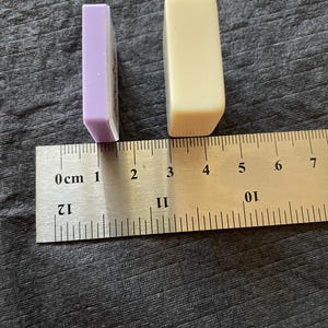 May include: Two rectangular objects, one lavender and one cream, are positioned next to a ruler. The lavender object is approximately 1 inch long. The cream object is approximately 1.5 inches long. The objects are on a textured, dark gray surface.
