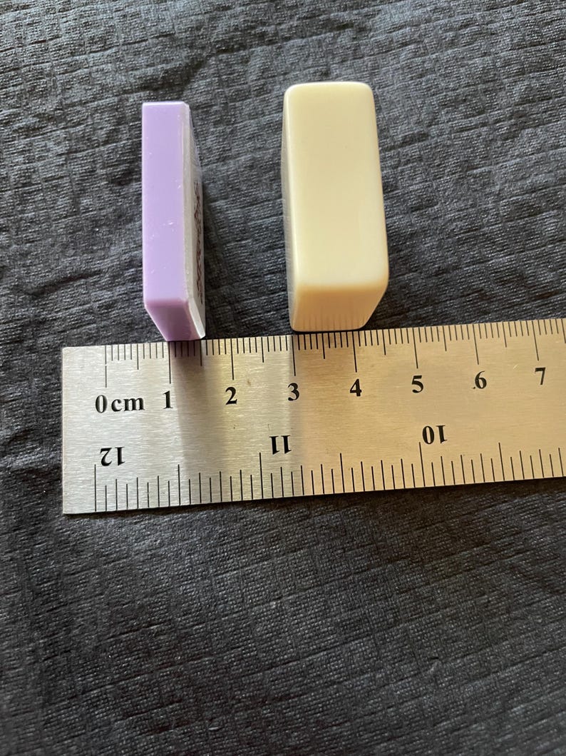 May include: Two rectangular objects, one lavender and one cream, are positioned next to a ruler. The lavender object is approximately 1 inch long. The cream object is approximately 1.5 inches long. The objects are on a textured, dark gray surface.