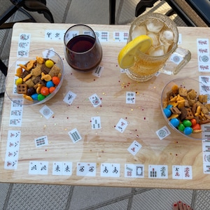 May include: A rectangular light wood serving tray featuring a Mahjong tile pattern printed along the edges and scattered across the surface. Two clear bowls filled with snack mix, a glass of dark liquid, and a glass mug with ice and lemon are present.