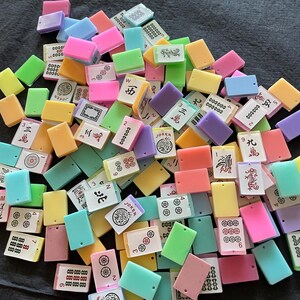 May include: A collection of colorful Mahjong tiles in various shades of pink, green, yellow, blue, and purple. The tiles feature Chinese characters and symbols, including numbers, circles, and dragons. The tiles are rectangular and made of a glossy material.