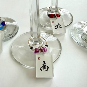 May include: Wine glass charms with rectangular white tags featuring black Chinese characters and the letters W, N, S, and E. The charms are adorned with colorful beads and attached to the wine glasses with gold rings.
