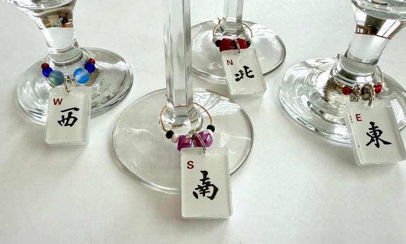 May include: Wine glass charms with rectangular white tags featuring black Chinese characters and the letters W, N, S, and E. The charms are adorned with colorful beads and attached to the wine glasses with gold rings.