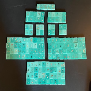 May include: A collection of turquoise Mahjong tiles arranged on a dark surface. The tiles feature gold and red decorative elements, including fish and floral designs. The tiles are rectangular and vary in size, suggesting a complete set for the game.
