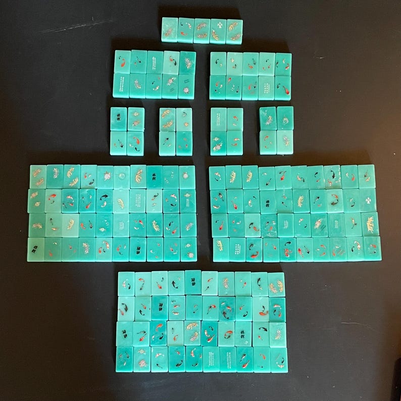 May include: A collection of turquoise Mahjong tiles arranged on a dark surface. The tiles feature gold and red decorative elements, including fish and floral designs. The tiles are rectangular and vary in size, suggesting a complete set for the game.
