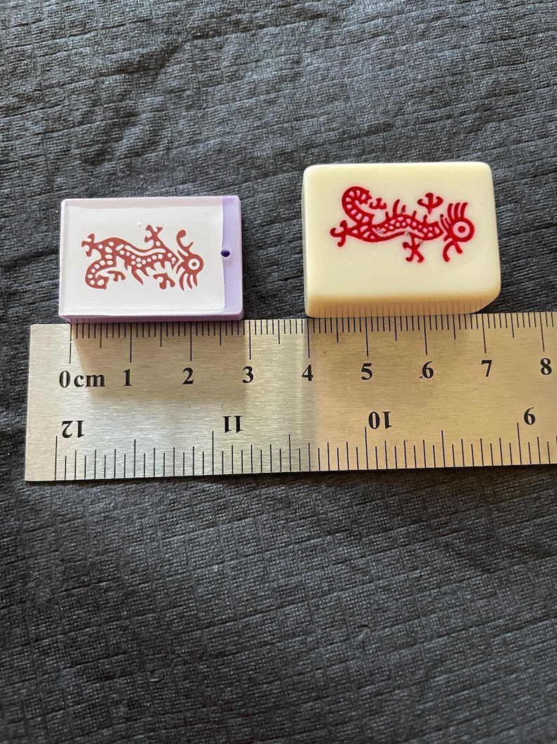 May include: Two Mahjong tiles with red dragon designs. One tile is rectangular with a purple border, the other is square and off-white. A ruler shows measurements in centimeters.