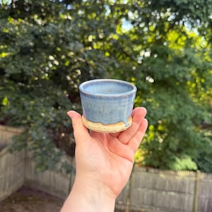 Könnte beinhalten: Eine handgefertigte Keramiktasse mit hellblauer Glasur und beigem Boden. Die Tasse wird in einer Hand vor dem Hintergrund von grünem Laub und einem Holzzaun gehalten. Die Tasse ist etwa 7.6 cm hoch.