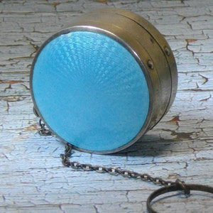 May include: A round, silver compact with a light blue enamel top and a chain attached. The compact has a decorative pattern on the top.