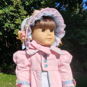 May include: A doll wearing a pink coat with blue buttons and trim over a white dress with small blue dots. The doll has a matching pink bonnet with blue lace trim and floral accents, and holds a small pink and white beaded accessory in one hand.