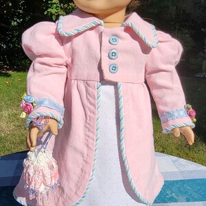 May include: A doll with brown hair wearing a light pink coat featuring blue buttons and blue and white twisted cord trim. The coat is worn over a white dress with small pink polka dots. The doll holds a small, beaded, floral-embellished pouch.
