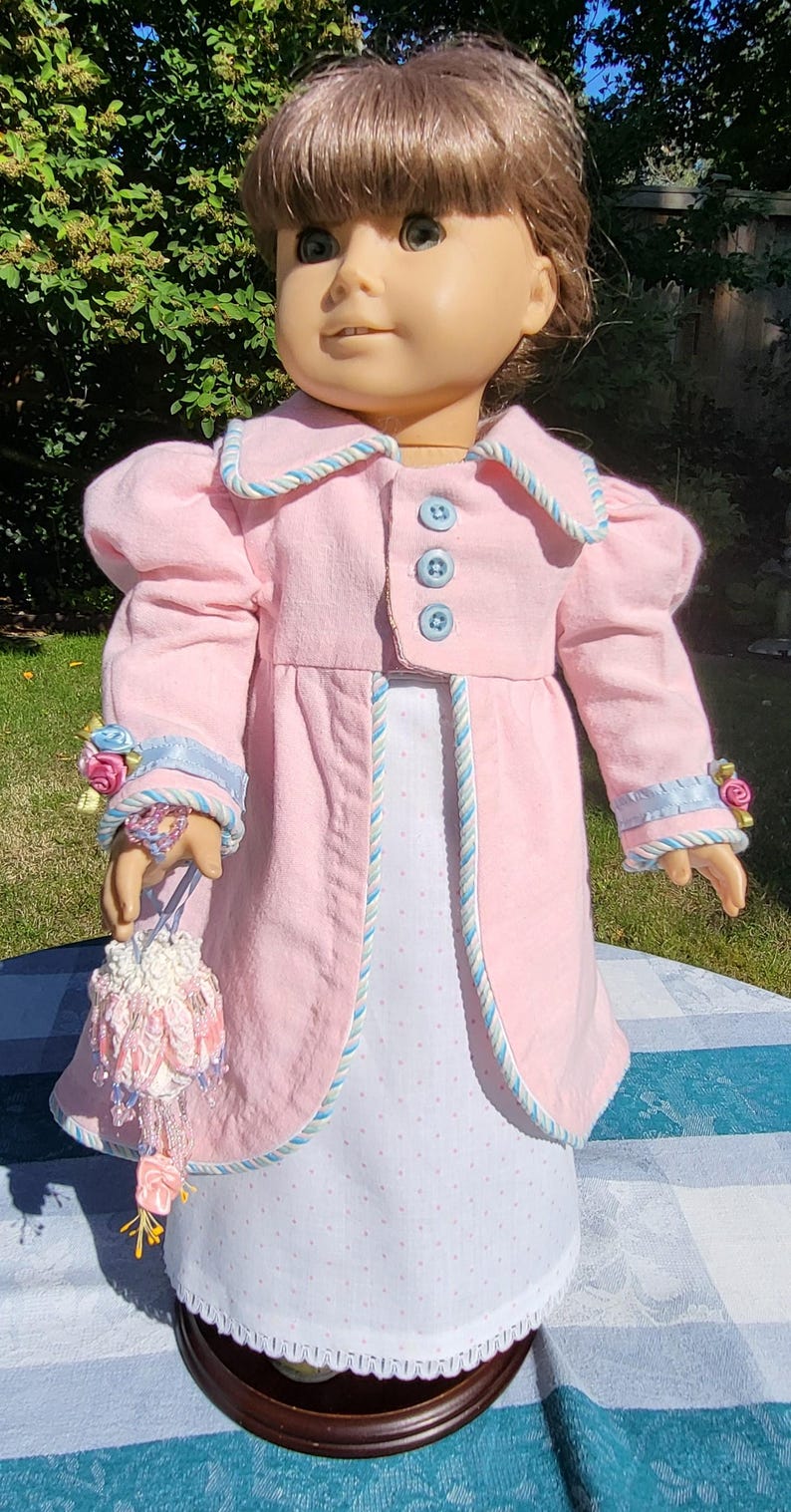 May include: A doll with brown hair wearing a light pink coat featuring blue buttons and blue and white twisted cord trim. The coat is worn over a white dress with small pink polka dots. The doll holds a small, beaded, floral-embellished pouch.