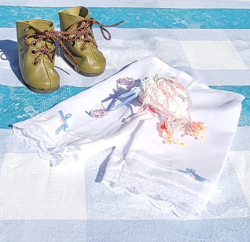 May include: A pair of olive green leather boots with brown patterned laces and a set of white fabric bloomers featuring delicate lace trim, light blue ribbon accents, and a small decorative floral embellishment with pink and yellow petals.