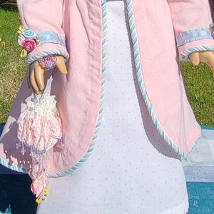 May include: A light pink coat with blue and white striped trim on the cuffs and edges, worn over a white dress with small pink polka dots. A small, ornate white crocheted drawstring pouch with pink ribbon accents and hanging beads is held.