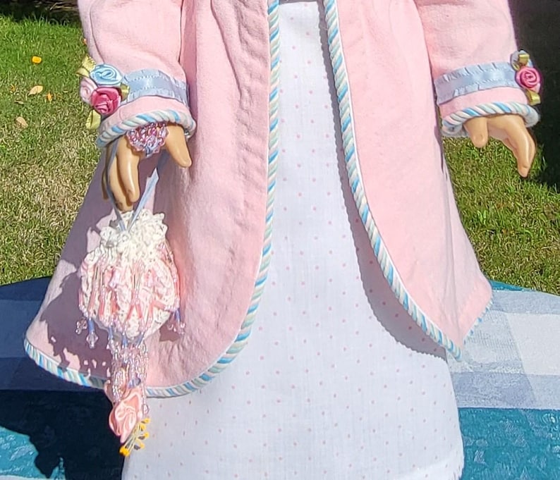 May include: A light pink coat with blue and white striped trim on the cuffs and edges, worn over a white dress with small pink polka dots. A small, ornate white crocheted drawstring pouch with pink ribbon accents and hanging beads is held.