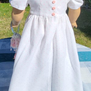 May include: A white dress with small pink polka dots, featuring short puff sleeves, a square neckline with delicate lace trim, and a vertical row of four small pink buttons down the centre of the bodice, paired with a small beaded drawstring bag.